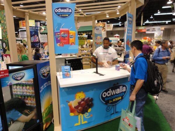 An Odwalla Smoothies for Kids booth with a blue counter and banners. A man behind the counter serves a visitor with a backpack, and a cooler stands at left.