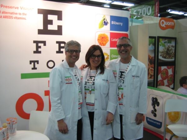 Two men and a woman in white lab coats and glasses stand together in front of an eye chart display at the OJO booth. A Quorn sign appears in the background.