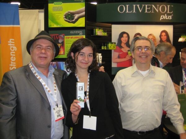 Two men and a woman stand in front of an Olivenol Plus sign; the woman holds up a small product box, and other people are visible behind them.