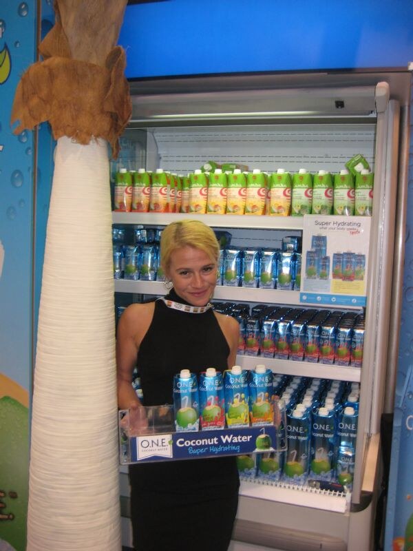 A blonde woman in a black dress holds a tray of O.N.E. Coconut Water cartons in front of a cooler stocked with O.N.E. and Jumba cartons. A palm trunk stands at left.