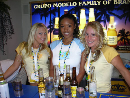 Three smiling women in yellow and white tops and bead necklaces stand behind a bar with Modelo beer bottles and cups, beneath a Grupo Modelo Family of Brands sign.