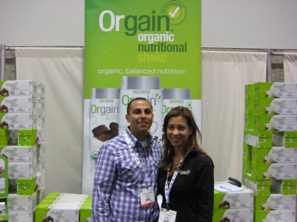 A smiling man in a plaid shirt and a woman in a black jacket stand in front of an Orgain Organic Nutritional Shake banner, flanked by stacked green Orgain shake cases.