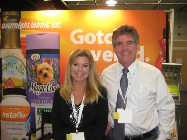 A smiling blonde woman and a man in a white shirt and tie stand in front of an Overnight Labels, Inc. backdrop with sample Four Paws Magic Coat and Instaflex bottles.