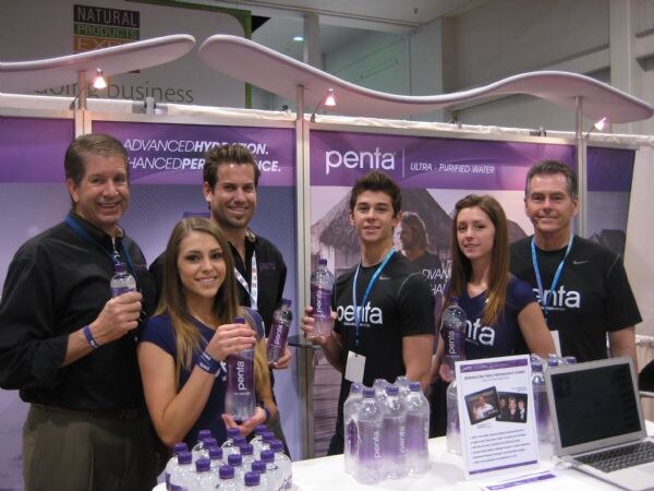 Six people in dark Penta shirts, some holding Penta water bottles, pose at a booth with purple Penta banners. Bottles and a laptop sit on the table in front of them.