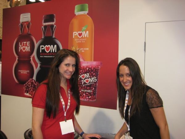 Two smiling women with long dark hair stand in front of a red POM Wonderful banner showing Lite POM and POM Wonderful bottles, a POM tea bottle and a cup of POM Poms.
