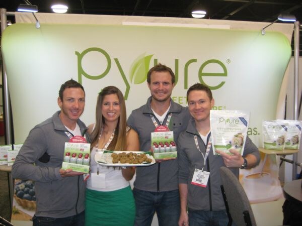 Three men and a woman pose in front of a green Pyure banner, holding Pyure stevia products and a plate of snacks.