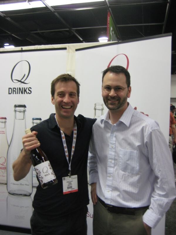 Two men pose in front of a Q Drinks banner showing tall bottles. The man on the left holds a bottle with a Q label.