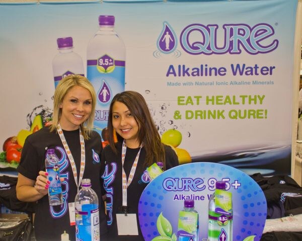 Two smiling women in black Qure shirts hold bottles of Qure Alkaline Water in front of a banner reading Eat Healthy & Drink Qure, with a Qure 9.5+ display sign.