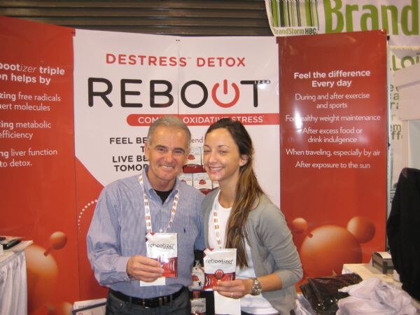 A smiling man and a smiling woman each hold a Rebootizer package in front of a red Reboot banner reading Destress Detox.
