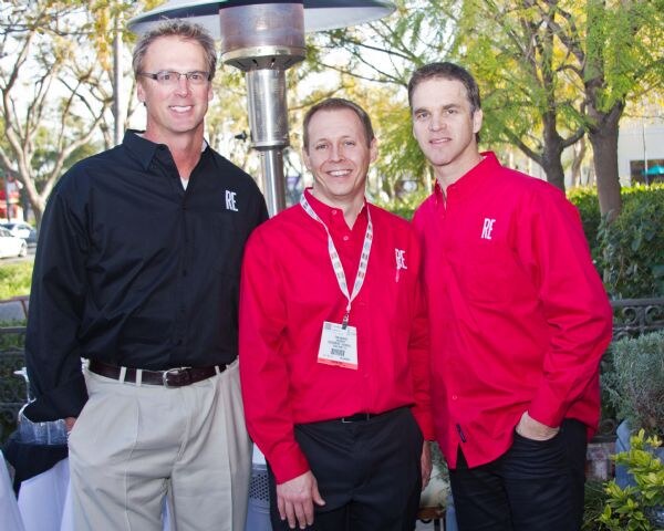 Three men stand outdoors near a patio heater, wearing black and red button-up shirts with an RE logo. The man in the middle wears a badge on a lanyard.