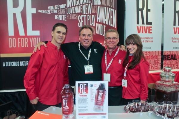 Four people in red and black shirts pose with arms around each other at the RE booth, with a RE Vitalize bottle and sign on the table and a glass dispenser with cups at right.
