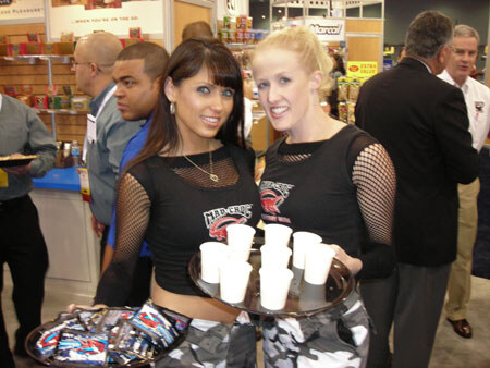 Two women in black Hard Rock mesh-sleeved tops hold trays of small white cups and packaged items in a crowded trade show aisle, with several men standing behind them.