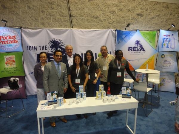 Seven people stand behind a white table with boxed products in a booth with Reya Coconut Water and Pocket Pretzels banners on the walls.