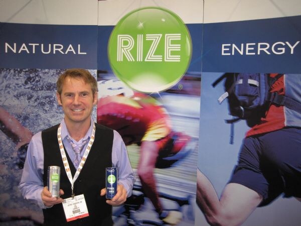 A smiling man in a dark vest and lanyard holds two small Rize cans in front of a Rize Natural Energy banner showing a cyclist and a runner.