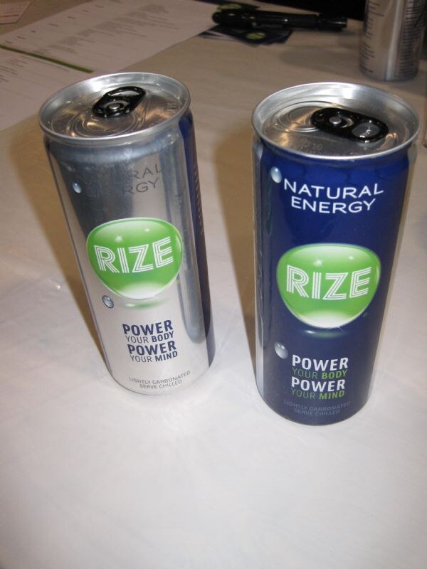 Two tall Rize Natural Energy cans, one silver and one dark blue, stand on a white tablecloth, each with a green Rize logo and the text Power Your Body Power Your Mind.