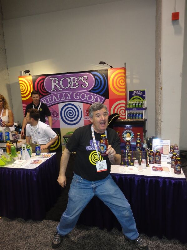 A man in a black Rob's Really Good shirt holds a bottle and strikes a playful pose in front of the colorful Rob's Really Good booth, where other people stand behind the table.