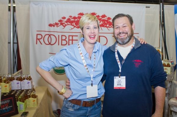A woman in a light blue shirt and a bearded man in a navy sweatshirt smile with arms around each other in front of a Rooibee Red banner, with bottles on a table at left.