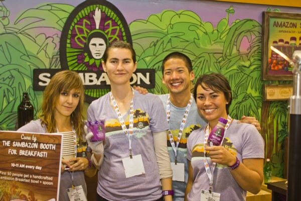 Four smiling people in matching gray Sambazon T-shirts stand together at the booth, two holding small purple bottles, beneath a Sambazon logo and a jungle mural.