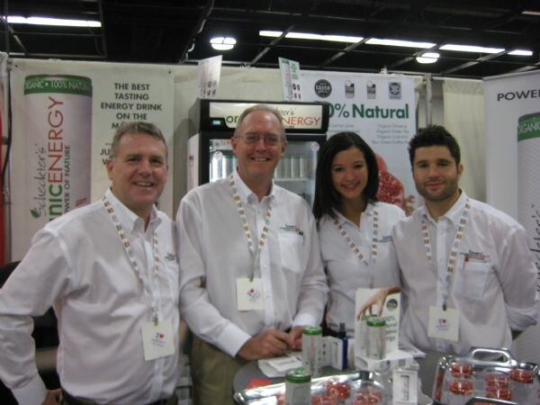 Four people in white shirts and lanyards, three men and a woman, stand together at the Scheckter's Organic Energy booth with cans and a tray of small cups on the table.