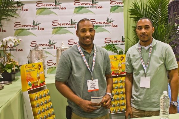 Two men in green polo shirts and lanyards stand at the Sereni Tea booth, with Sereni Tea banners behind them and boxed product displays on either side.