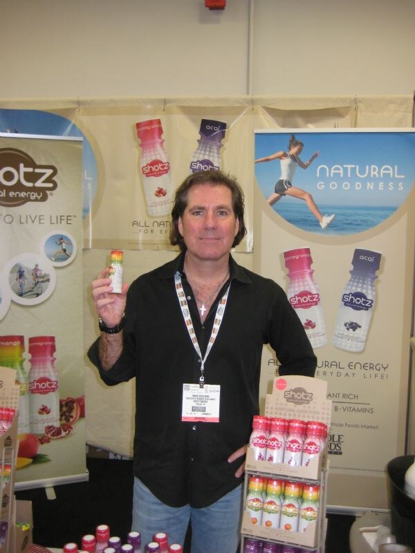 A man in a black shirt and lanyard holds up a small Shotz bottle in front of Shotz banners, with a display of Shotz bottles on the table beside him.