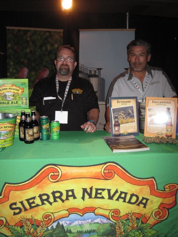 Two men sit behind a green Sierra Nevada table with beer bottles, cans, a Pale Ale carton and Ruthless Rye IPA and Kellerweis signs. The Sierra Nevada logo is on the table front.