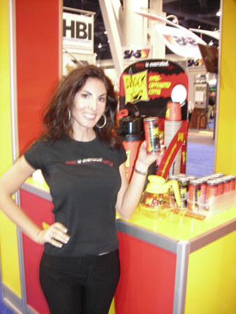 A smiling woman with dark wavy hair in a black T-shirt holds an energy drink can at a yellow and red counter with cups and cans, near HBI International and SAS signs.