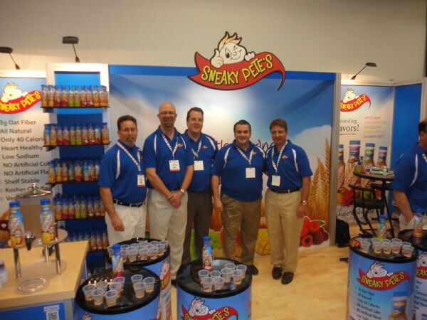 Five men in blue polo shirts and lanyards stand in front of a Sneaky Pete's booth with bottle shelves, sample cups and a sign with the brand's cartoon logo.