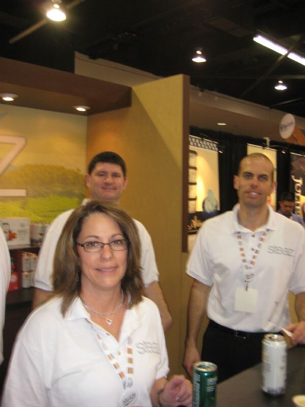A woman with glasses and two men, all in white Steaz polo shirts and lanyards, stand in a booth. Steaz cans are in the foreground.