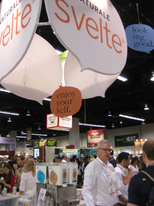 Hanging Svelte signs with slogans like "enjoy your self" hang over a beverage dispenser. A man in a white shirt stands in front, with Organic Valley and Organic Prairie signs behind.