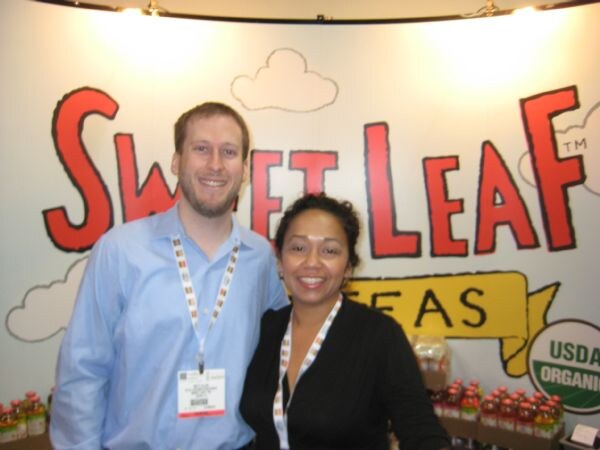 A smiling man in a light blue shirt and a smiling woman in black stand in front of a Sweet Leaf Tea backdrop with bottles on a shelf at the lower right.