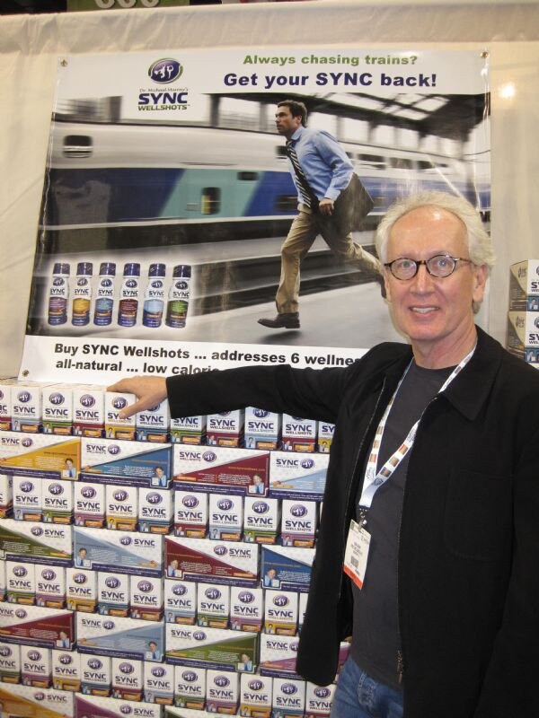 A gray-haired man in glasses and a black jacket points to a stacked display of Sync Wellshots boxes beneath a banner reading "Get your SYNC back!"