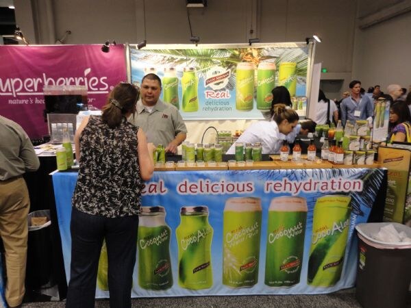 A woman faces a booth counter where a man stands behind a Taste Nirvana coconut water banner. A Superberries sign is at left.