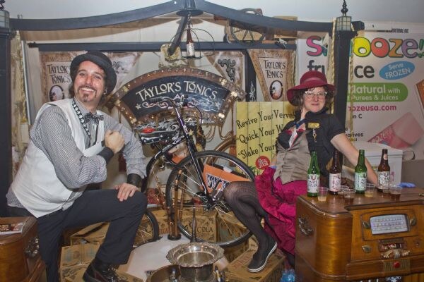 A man in a vest and hat and a woman in a red hat sit by a bicycle in a steampunk-style Taylor's Tonics booth with bottles on an old radio cabinet. A Zeus sign is behind.