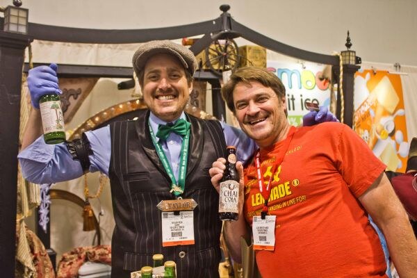 Two smiling men in blue gloves hold bottles, one labeled Chai Cola. One wears a green bow tie and cap, the other an orange Lemonade shirt.