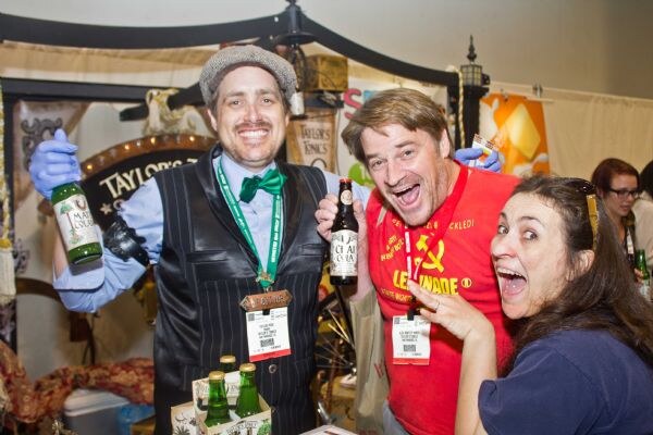 Three people laugh at a Taylor's Tonics booth. One in a vest and green bow tie holds a bottle, another in a red shirt holds a bottle, and a woman leans in with her mouth open.