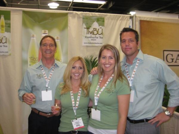 Two men and two women in green shirts and lanyards pose together in front of TheBü Kombucha Tea banners showing bottles.