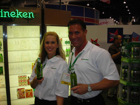 A smiling woman and man in white Heineken polo shirts each hold a green Heineken bottle in front of lit display coolers and a partial Heineken sign.