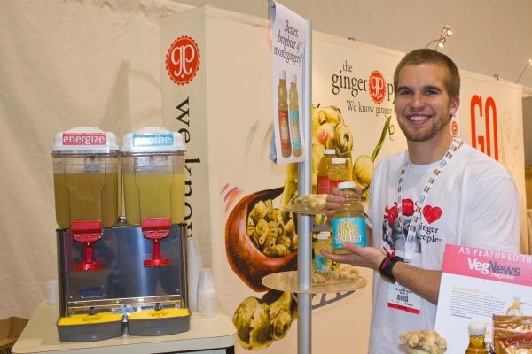 A smiling man holds a Ginger Souther bottle beside a Ginger People banner and two beverage dispensers. A VegNews magazine feature sign is at the lower right.