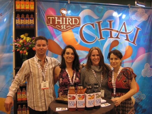Two men and women... one man and three women stand behind a table of Third St Chai iced tea bottles in front of a colorful Third St Chai banner.