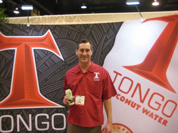 A man in a red Tongo polo holds a bottle of coconut water in front of large red and black Tongo Coconut Water banners.