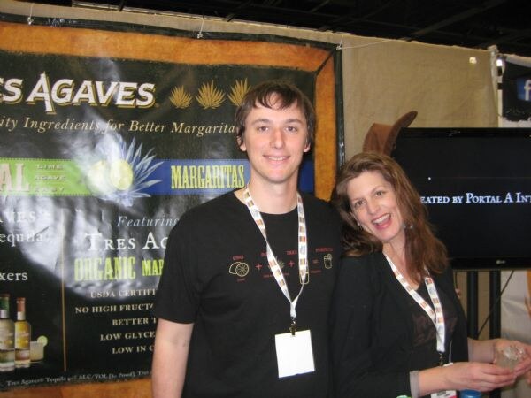 A young man in a black shirt and a woman in a dark jacket smile beside a Tres Agaves banner about margarita mixers, with bottles at the lower left.