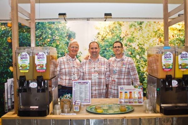 Three men in plaid shirts stand behind a counter at an Uncle Matt's booth, between juice dispensers, with an orange grove backdrop and juice bottles on display.