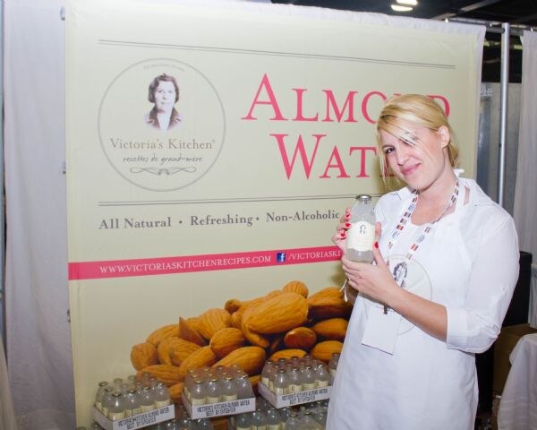 A woman in a white coat holds a bottle of almond water in front of a Victoria's Kitchen banner with almonds and cases of bottles below.