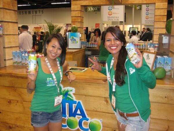 Two smiling women in green Vita Coco clothing hold cartons of coconut water at a wooden Vita Coco booth. A Manuka Doctor sign is in the background.