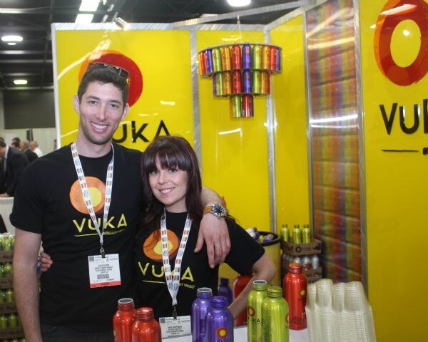 A man and a woman in black Vuka shirts pose with arms around each other in front of yellow Vuka displays, with colorful bottles on the table below.