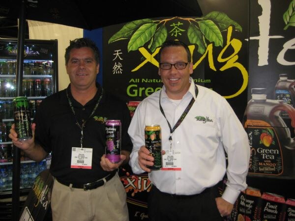 Two smiling men wear lanyards and hold canned drinks in front of a green Xing tea display; one is in a black polo, the other in a white shirt and glasses.