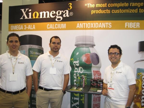 Three men in white polo shirts and lanyards stand at a table of boxed products in front of a Xiomega banner showing a bottle and the words Omega 3-ALA, Calcium, Antioxidants and Fiber.
