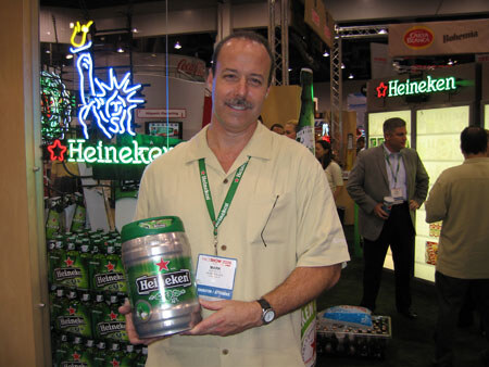 A smiling man with a mustache and lanyard holds a Heineken mini keg in front of stacked Heineken cases and a neon Heineken sign with a Statue of Liberty figure.