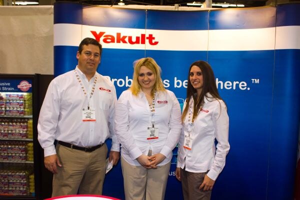 A man and two women in white shirts and lanyards stand together in front of a blue Yakult banner, with a refrigerated case of products on the left.